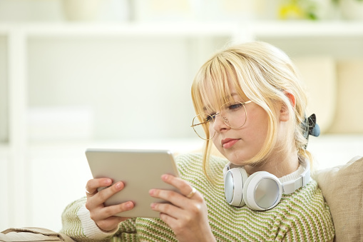 Side view of smiling teenage girl sitting on sofa in the living room at home, holding iPad in hands.