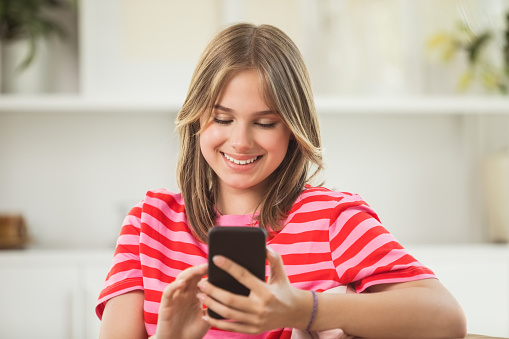 Front view of smiling teenage girl sitting on sofa in the living room at home, holding mobile phone in hands.