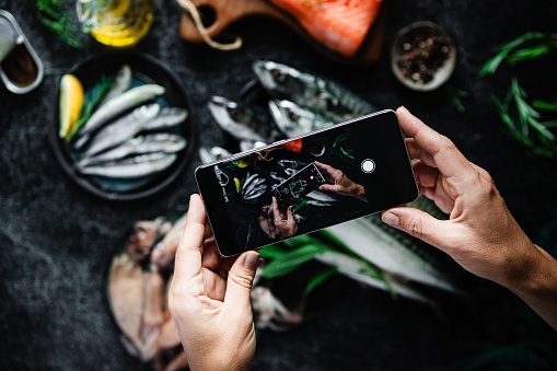 Close-up of woman photographing healthy omega 3 rich food with her mobile phone on kitchen table. Point of view of a woman hand holding smartphone and taking photos of different fishes, spices and food ingredients on a black table.