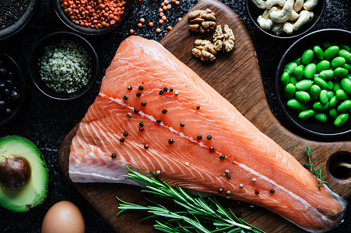 Top view of omega 3 rich food ingredients on table. Close-up of fresh raw salmon steak with black pepper on wooden cutting board, rosemary, egg, avocado, black salt, lentils, walnut, cashew nut on kitchen table.