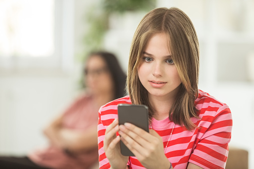 Front view of worried teenage girl sitting on sofa in the living room at home, holding mobile phone in hands with her defocused mother in the background.