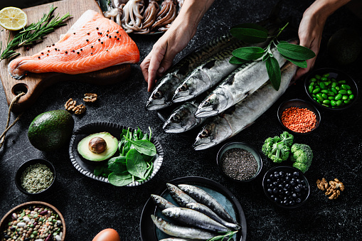 Female hands arrange food ingredients with rich omega-3 and healthy fats. Cropped shot woman placing foods high in fatty acids including vegetables, seafood, nut and seeds on black table.