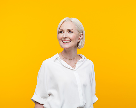 Happy blond hair mature woman wearing white shirt standing against yellow background and looking away. Studio shot.