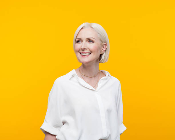 Portrait of beautiful mature woman Happy blond hair mature woman wearing white shirt standing against yellow background and looking away. Studio shot. older blonde woman stock pictures, royalty-free photos & images