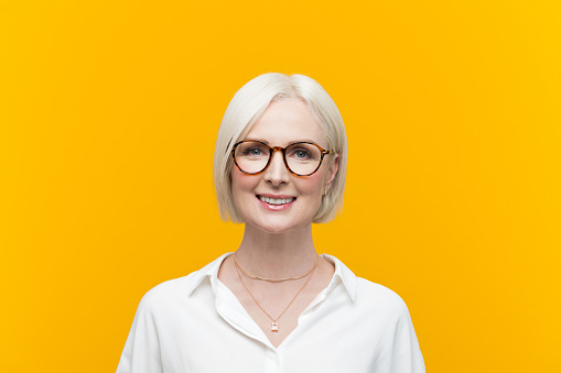 Happy blond hair mature businesswoman wearing white shirt standing against yellow background and smiling at camera. Studio shot.