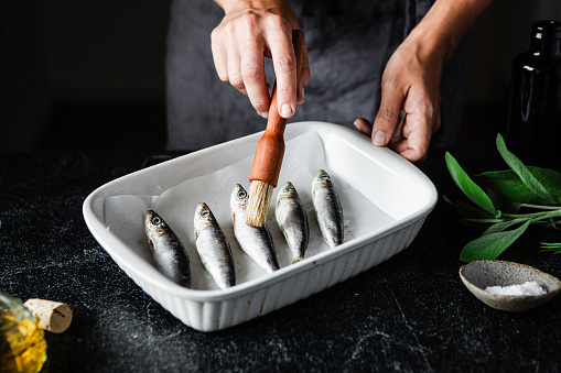 Close-up of female chef hands applying oil on raw sardines on a white tray. Close-up of preparing healthy and delicious seafood in kitchen.