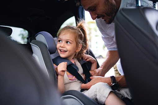 A smiling Caucasian male preparing his adorable child (looking away) for driving in a vehicle.