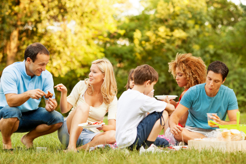 Two families with children, sitting on grass and enjoy barbecue meal outdoors in nature.