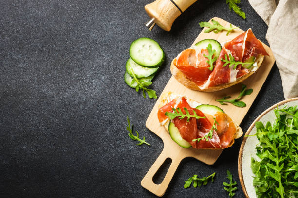 istock Open sandwiches with cream cheese, prosciutto, cucumber and arugula at black table. 1644793565