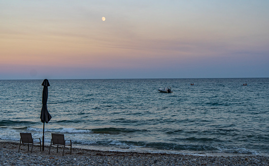 Sunset on the beach with two chairs and umbrellas during sunset with moon on the beach of the Aegean sea at the end of the beach season in Greece