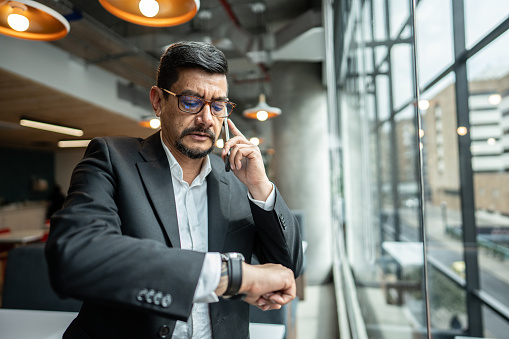 Mature man checking the time on his watch while talking on mobile phone in the office