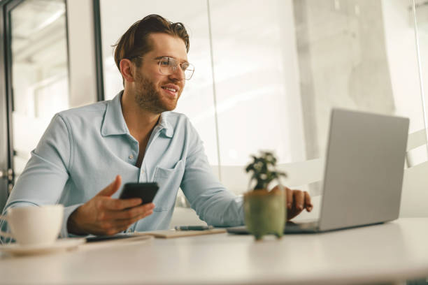 istock Smiling male entrepreneur holding phone while working on laptop in modern office 1644977632