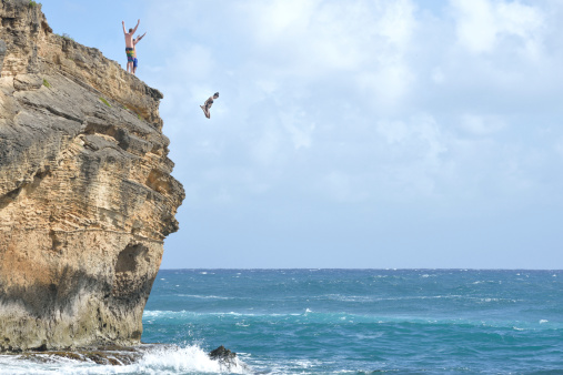 A young woman in her 20s is cliff diving in Kauai while her friends cheer her on.