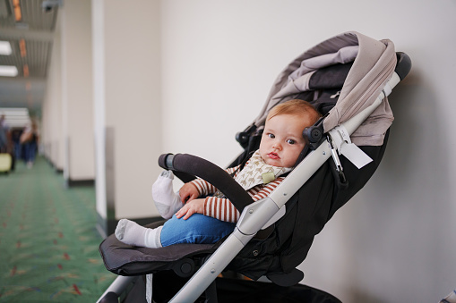 9 month old boy of Eurasian, Hawaiian and Pacific Islander ethnicities sits patiently in a stroller by the wall of an airport.