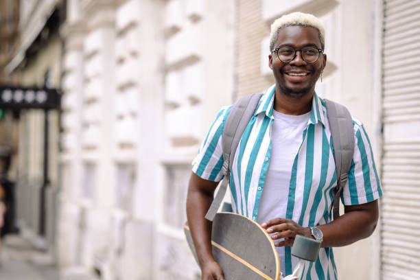 Portrait of urban African American man with longboard in the city Young man carrying a longboard and backpack. He is smiling and looking at the camera black man blonde hair stock pictures, royalty-free photos & images