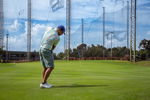 Multiracial, male golf professional wearing casual clothing, practices his swing at a driving range on a sunny, warm summer day.