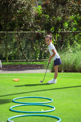 Adorable, active young girl of Asian ethnicities watches the golf ball she just hit go into rings laid on the ground as she practices.
