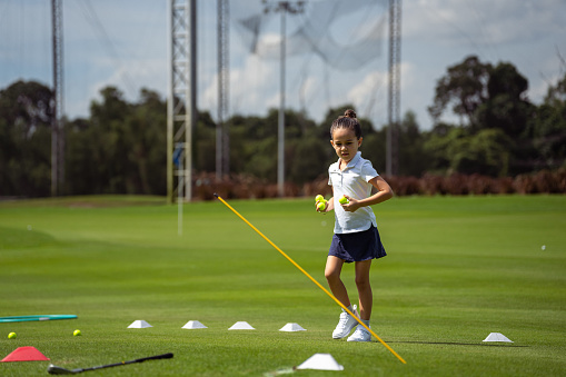 Kindergarten aged girl of Asian ethnicities jogs along a golf course, picking up balls after practicing on a sinner, summer day.