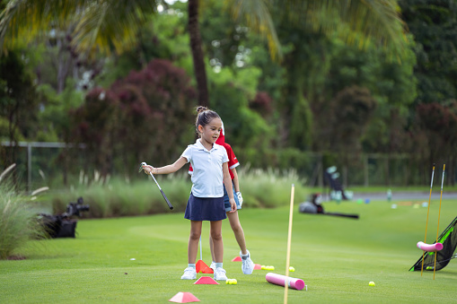 Kindergarten aged girl of Asian ethnicity stands, holding her golf club as she follows a circuit of golf drills to improve her swing.