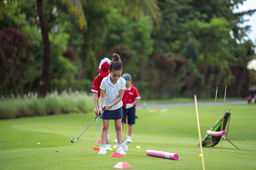 Kindergarten aged girl of Asian ethnicity practices her golf swing by participating in drills set up on a golf course on a sunny, summer day.