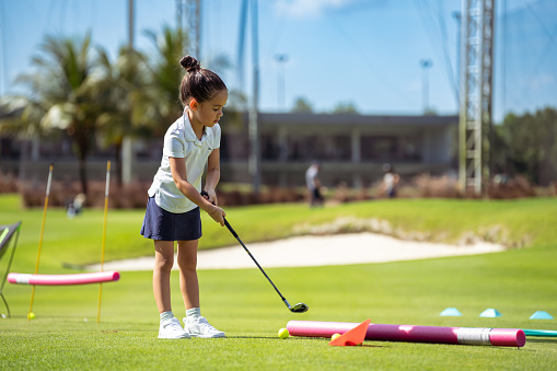 Kindergarten aged girl of Asian ethnicity practices her golf swing by participating in drills set up on a golf course on a sunny, summer day.