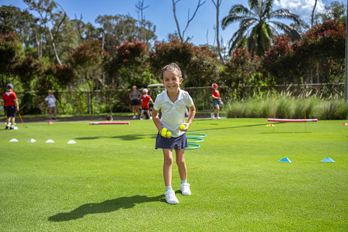 Adorable, active young girl of Asian ethnicities smiles and giggles at the camera as she picks up golf balls off a golf course.