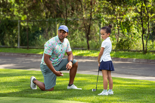 Loving, active father of Asian ethnicity kneels beside his kindergarten aged daughter, showing her how to swing a golf club as they spend time on a course.