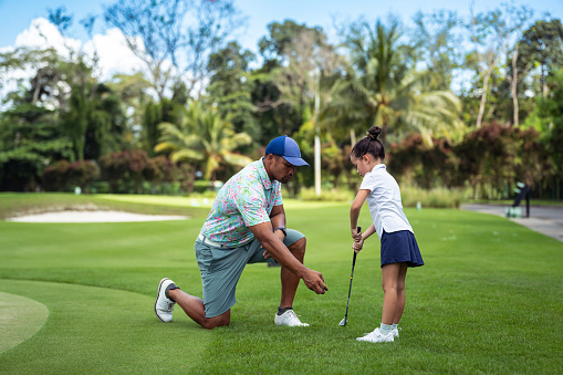 Loving, active father of Asian ethnicity kneels beside his kindergarten aged daughter, showing her how to swing a golf club as they spend time on a course.