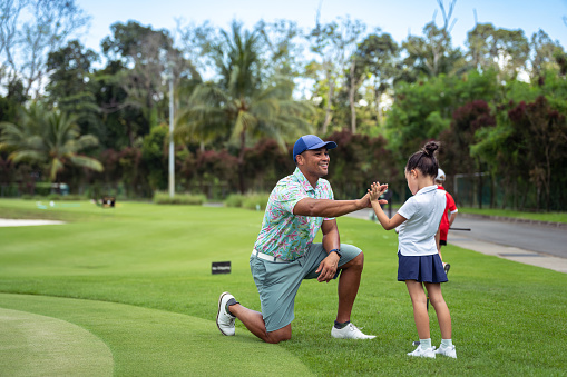 A kindergarten aged girl of Asian ethnicities stands beside her proud, multiracial, male golf coach as they high five over her accomplishments on the golf course.