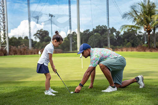 A loving, multiracial father kneels on the golf course, repositioning a golfball as he instructs his kindergarten aged daughter on how to putt.