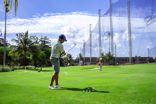 Multiracial, male golf professional wearing casual clothing, practices his swing at a driving range on a sunny, warm summer day.