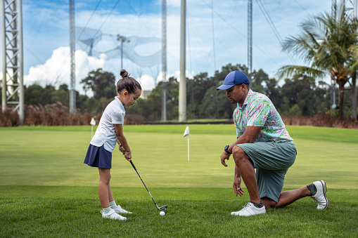 A multiracial, male professional golf instructor kneels on the course as he instructs a kindergarten aged girl of Asian ethnicity how to putt a golfball.
