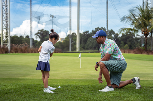 A multiracial, male professional golf instructor kneels on the course as he instructs a kindergarten aged girl of Asian ethnicity how to putt a golfball.