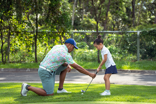 Male, multiracial golf professional kneels next to a kindergarten aged girl of Asian ethnicities, touching her golf club as he coaches her on her swing.
