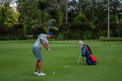 Young man of multiracial ethnicity stands on a golf course with his bag of clubs, practicing his swing on a summer day.