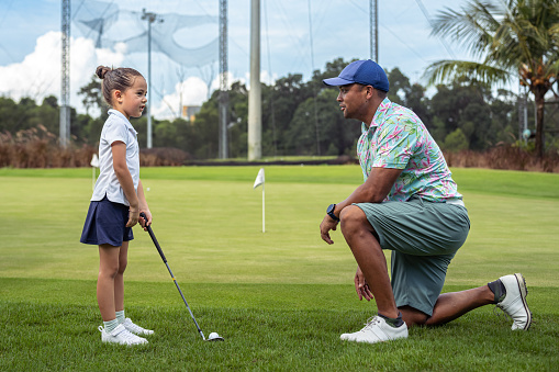 Loving and active multiracial father kneels on a golf course as he instructs his kindergarten aged daughter how to putt a golfball.
