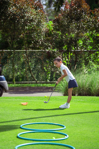 Adorable, active young girl of Asian ethnicities watches the golf ball she just hit go into rings laid on the ground as she practices.