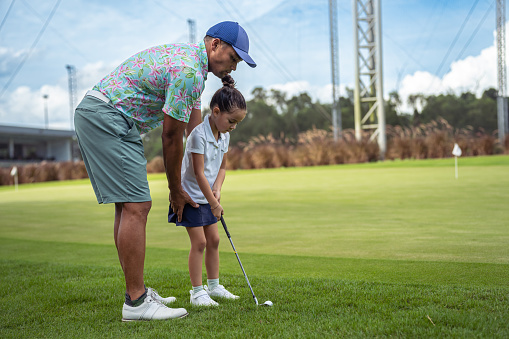 Kindergarten aged girl of Asian ethnicity is being shown how to correctly swing a golf club by her loving, active father on a golf course.