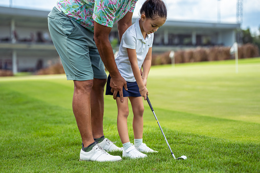 Kindergarten aged girl of Asian ethnicity is being shown how to correctly swing a golf club by a professional on a golf course.