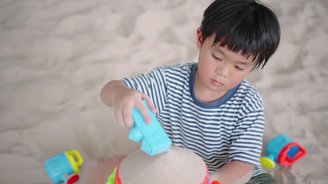 A little boy is pouring sand through the funnel and making the wheel spin in the sandpit alone.