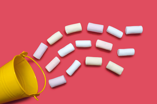 A small yellow bucket and a lot of scattered multi-colored marshmallows on a pink background, top view. Confectionery concept of marshmallows in a worm of water that flows from a bucket