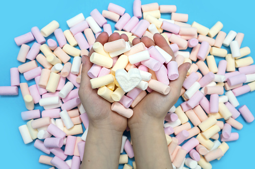 A lot of multi-colored air marshmallows on a blue background and a child's hands close-up. Favorite sweet dessert for kids or topping hot drinks in the form of cocoa or hot chocolate