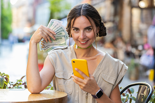 Happy rich woman counting money dollar cash, use smartphone calculator app, plans to order food drink delivery online, booking hotel room. Pretty girl tourist sits at table in city cafeteria outdoors