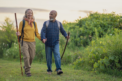 A senior couple hike together on a warm summers evening.  They are dressed casually, wearing backpacks and have walking sticks in hand for stability.