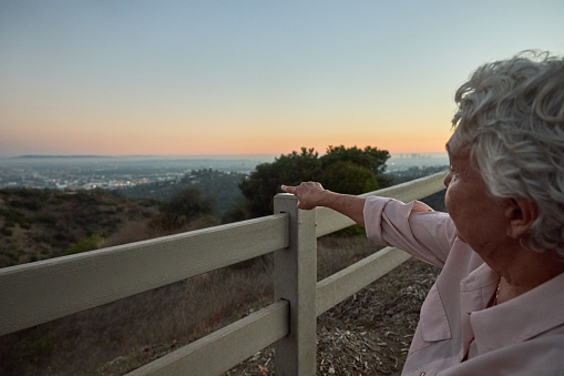 Senior woman with gray hair, sitting at a viewing area above the city of Los Angeles, at sunset.