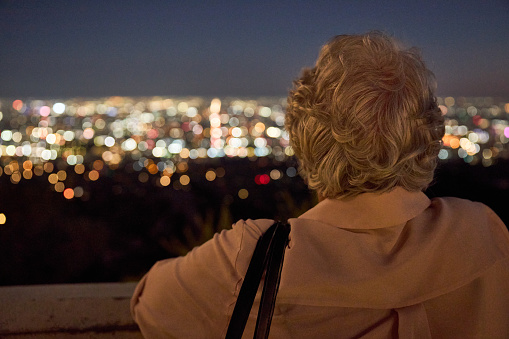 Back of a gray-haired lady looking at Los Angeles lights from an elevated area.