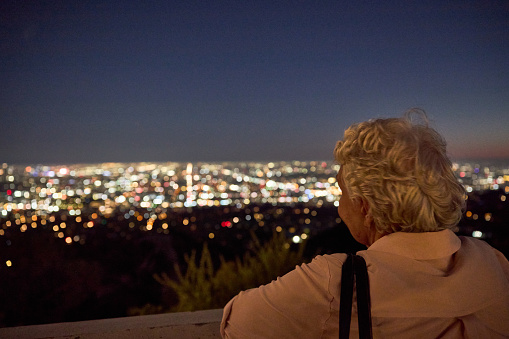 Back of a gray-haired lady looking at Los Angeles lights from an elevated area.