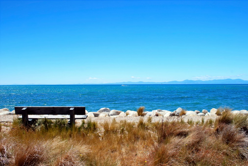 Looking out through the tussock grass to an empty seat and the turquoise waters of the Tasman Sea. Taken in Marahau, the gateway to the Abel Tasman Nation Park in New Zealand's South Island.