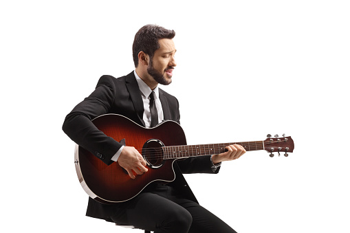 Musician with an acoustic guitar sitting on a chair and playing isolated on white background