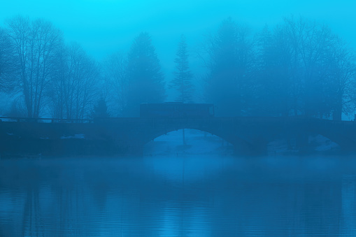Tourist shuttle bus crossing the old stone bridge at Bohinj lake in foggy winter morning, blue toned image